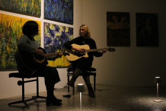 Recital Małgorzaty Karpińskiej i Aleksandry Górnej. Kobiety siedzą na taboretach, grają na gitarach, patrzą na siebie. Na ścianie wyeksponowane prace.