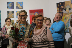 Uśmiechnięta Krystyna Ruminkiewicz pozuje do zdjęcia z jedną z uczestniczek wernisażu. Trzyma czerwone róże i upominkowa torbę z serduszkami i napisami Love. W tle goście i prace wyeksponowane na ścianach.