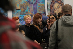 Grupa gości wernisażu. Po prawej plecy mężczyzny w szarej bluzie z kapturem. W centrum kadru Renata Rudowicz z CKiS w Koninie z aparatem.