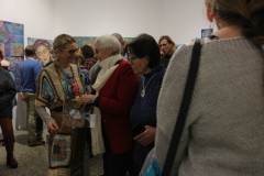 Tłum gości. Krystyna Ruminkiewicz w trakcie rozmowy z dwiema uczestniczkami. Pół kadru zajmują plecy kobiety stojącej po prawo. Ubrana jest w szary sweter, Przez ramię przewieszony ciemny pasek torebki.