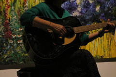 Kobieta w ciemnej spódnicy i zielonej bluzce gra na gitarze. Za nią prace - namalowane kwiaty.