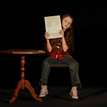 Dziewczynka w dżinsach siedzi na krześle. Unosi w górę narysowany portret. Obok niej stolik.