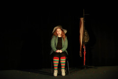 Dziewczynka w kapeluszu, zielonej bluzie i pasiastych rajstopach siedzi na krześle na scenie.  Za nią wieszak.