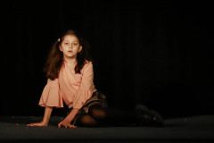 Dziewczynka opierając się na dłoniach leży na scenie. Włosy związane w dwie kitki, Bluzka w kolorze pudrowym.