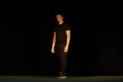 Na scenie chłopak w ciemnych spodniach, ciemnej koszulce i sportowych butach. Za nim mrok sceny.