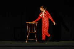 Dziewczyna w czerwonych piżamach w serduszka. Na szyi kokarda. Prawą dłonią chwyta poręcz drewnianego krzesła.