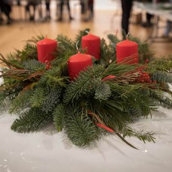 Na stole przykrytym folią stoi wieniec zrobiony z gałązek iglaków. Ozdobiony czterema czerwonymi świecami.