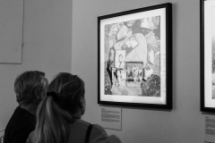 Mężczyzna i kobieta stoją tyłem do obiektywu. Oglądają zawieszoną na ścianie fotografię.  Przedstawia ona kilka zdjęć ułożonych na dużym kamieniu.