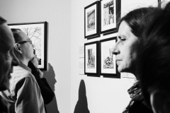 Na pierwszym planie twarze kobiety i mężczyzny. W tle  uczestnik wystawy przygląda się serii czterech fotografii zawieszonych na ścianie.