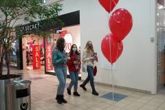 Trzy wolontariuszki z puszkami idą przez galerię. W tle sklep Top Secret. Na pierwszym planie trzy czerwone balony.