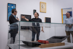 Na pierwszym planie przeszklona gablota z pracą. Za nią przy ścianie od lewej: Robert Brzęcki, gastykulujący Piotr Lutyński i Michał Kruszona.