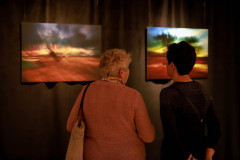 Sfotografowane tyłem dwie kobiety oglądają dwie prace zawieszone na ciemnej kotarze.
