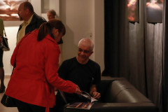Tadeusz Topolski siedzi na ciemnej kanapie. Obok niego stoi kobieta w czerwonym krótkim płaszczu. W ręce trzyma katalog.