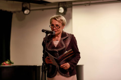 Krystyna Mikołajczak przy mikrofonie w planie amerykańskim. W dłoniach trzyma katalog.