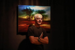 Tadeusz Topolski pozuje do zdjęcie. Ręce splótł przed sobą, ściskając katalaog. Za nim wisi jedna z prac. Pejzaż z dwoma drzewami.