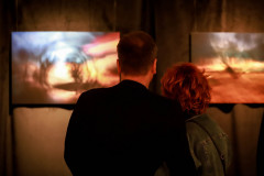 Stojący plecami do obiektywu kobieta i mężczyzna oglądają prace zawieszone na ciemnej kotarze.