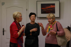 Trzy kobiety w trakcie rozmowy. W dłoniach trzymają szklanki lub kieliszki. Na ścianie w czarnej ramce  tytuł wystawy: Miraże Tadeusza Topolskiego.