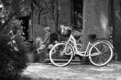 Biały rower oparty o pień drzewa.  W tle dwóch chłopców siedzi na plastikowym koniku. Zdjęcie czarno-białe.