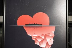 Plakat Titanic. Na czarnym tle czerwone serce przechodzące w górę lodową i czarny kształt transatlantyku.