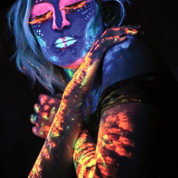 Popiersie kobiety w wizażu farb UV: ręce i rzęsy mają kolor pomarańczowy, zamknięte powieki różowy, natomiast twarz i ramiona są niebiesko-białe.  Dłonie skrzyżowane na barkach.
