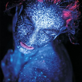 Portret kobiety w wizażu niebiesko-czerwonych farb UV.  Zdjęcie wykonane z góry. Modelka ma zamknięte powieki, uchylone usta i rozwichrzone włosy.