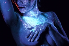 Kobieta w wizażu czerwono-niebieskich farb UV. Prawa ręka zgięta w łokciu dotyka czubka głowy. Dłoń lewej spoczywa na piersi.