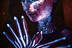 Kobieta w wizażu farb UV. Na dłoniach białe smugi pociągnięte wzdłuż kości.  Długie czarne włosy zaczesane na prawy bark.