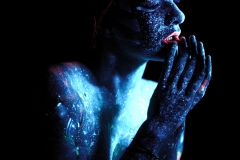 Popiersie kobiety w wizażu niebiesko-czerwonych farb UV. Skierowana jest bokiem do obiektywu. Siedzi w modlitewnej pocie: złączonymi dłońmi dotyka ust. Ma zamknięte powieki.