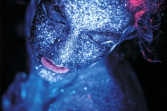 Portret kobiety w wizażu niebiesko-czerwonych farb UV.  Zdjęcie wykonane z góry. Modelka ma zamknięte powieki, uchylone usta i rozwichrzone włosy.