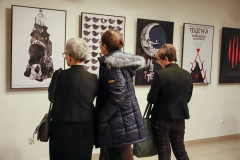 Trzy osoby stojące tyłem do obiektywu oglądają plakaty wyeksponowane na białej ścianie. Prace przedstawiają ptaki, księżyc oraz plakat do sztuki Witkacego Mątwa.