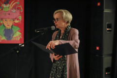 Krystyna Mikołajczak z CKiS w Koninie przy mikrofonie na statywie. W dłoniach trzyma teczkę. W tle ciemne kotary, fragment plakatu i głośniki.