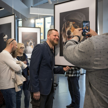 Błażej Zalasiński przybliża twarz do portretu owczarka collie w czapce. Po prawej ramię osoby robiącej zdjęcie telefonem. W tle mężczyzna i kobieta oraz wiszące prace.