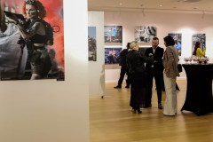 Po lewo na pierwszym planie kolorowa fotografia Darii Mach z bronią. W głębi goście podczas rozmówk, fragmenty prac i stolik z kieliszkami.