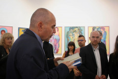 Sfotografowany z prawego profilu Sławomir Kuszczak trzyma dwie książki. W głębi uczestnicy wernisazu. Na białej ścianie rząd prac.