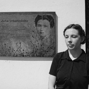 Młoda kobieta w czarnej koszuli z krótkimi rękawami. Siedzi niemal na wprost obiektywu. Po jej prawej stronie przymocowana do ściany tablica upamiętniająca Zofię Urbanowską.