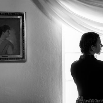 Po prawej stronie naprzeciw okna tyłem do obiektywu stoi kobieta w ciemnej koszuli z krótkimi rękawami. Twarz ma zwróconą w bok. Po lewej na białej ścianie zawieszony portret Zofii Urbanowskiej w podobnej pozycji.