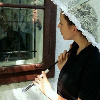 Kobieta w czarnej koszuli z krótkimi rękawami widziana od pasa w górę. W dłoniach trzyma opartą o prawy bark rączkę rozłożonego parasola. Spogląda w szybę okna, w której odbija się jej twarz.