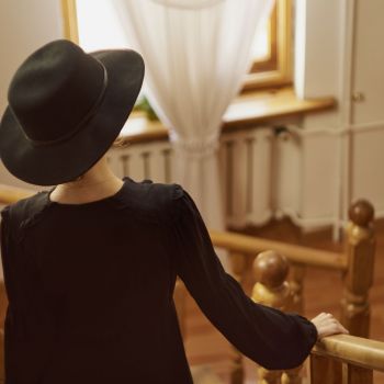 Kobieta w czarnej, zwiewnej sukience i czarnym okrągłym kapeluszu. Schodzi po schodach tyłem do obiektywu. W tle okno o drewnianych ramach, które przysłania biała zasłona.