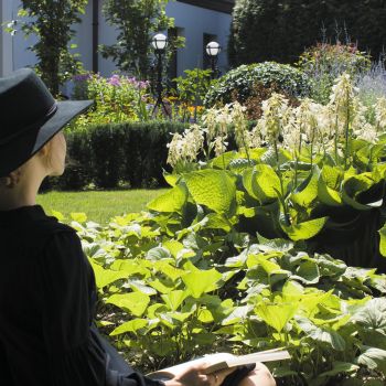 Po lewej stronie kobieta w czarnej, zwiewnej sukience i czarnym, okrągłym kapeluszu, spod którego wystaje fragment jasnego warkocza. Spogląda przed siebie. Po prawej bujna roślinność ogrodu. W tle białe ściany dworku.
