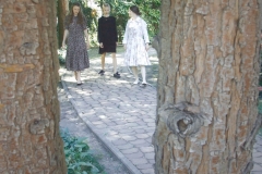 Trzy kobiety w zwiewnych sukienkach. Idą chodnikiem z kostki. Zdjęcie wykonane spomiędzy pni dwóch drzew.