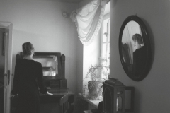 Kobieta w czarnej sukni stoi tyłem do obiektywu naprzeciwko drewnianego sekretarzyka z lustrem. Prawą dłoń trzyma na jego blacie. W zawieszonym na bocznej ścianie okrągłym lustrze odbija się jej postać. Zdjęcie czarno-białe.