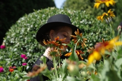 Młoda kobieta w czarnym, okrągłym kapeluszu stoi pośród kwiatów, które przysłaniają znaczą część jej sylwetki.