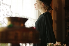 Młoda kobieta w czarnej, zwiewnej sukience i czarnym kapeluszu widziana bokiem stoi naprzeciw okna. Ma jasne włosy splecione w warkocz wystający spod rondla.  Na pierwszym planie zamglony klosz lampy.