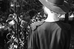 Szczupła kobieta w czarnej, zwiewnej sukience i czarnym, okrągłym kapeluszu. Stoi tyłem do obiektywu. Po lewej stronie zdobiona lampa ogrodowa. Zdjęcie czarno-białe.