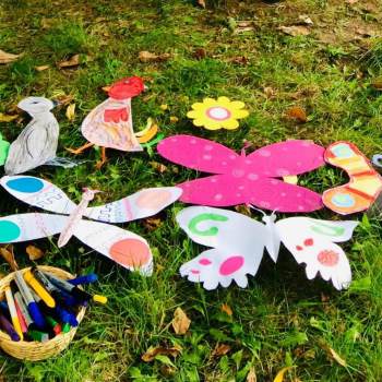 Na trawniku leżą wycięte z papieru i pokolorowane przez uczestników zajęć motyle i ptaki. Obok w pojemnikach znajdują sie kredki i pisaki.