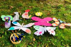 Na trawniku leżą wycięte z papieru i pokolorowane przez uczestników zajęć motyle i ptaki. Obok w pojemnikach znajdują sie kredki i pisaki.