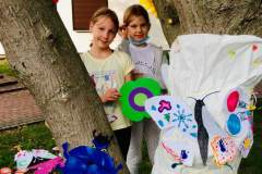 Między konarami drzewa dwie dziewczynki prezentują kwiat z kolorowego papieru.