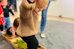 Dziecko trzymane za rękę, ubrane w beżowy sweter i czarne getry wchodzi do drewnianego pudełka z szyszkami.