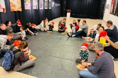 Uczestnicy zajęć - dorośli i dzieci - siedzą na ciemnej folii przykrywającej podłogę. Tworzą koło. Dzieci mają na głowach Mikołajkowe czapki.