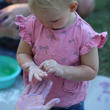 Dziewczynka w różowej koszulce zeskrobuje masę z dłoni. Głowa pochylona.